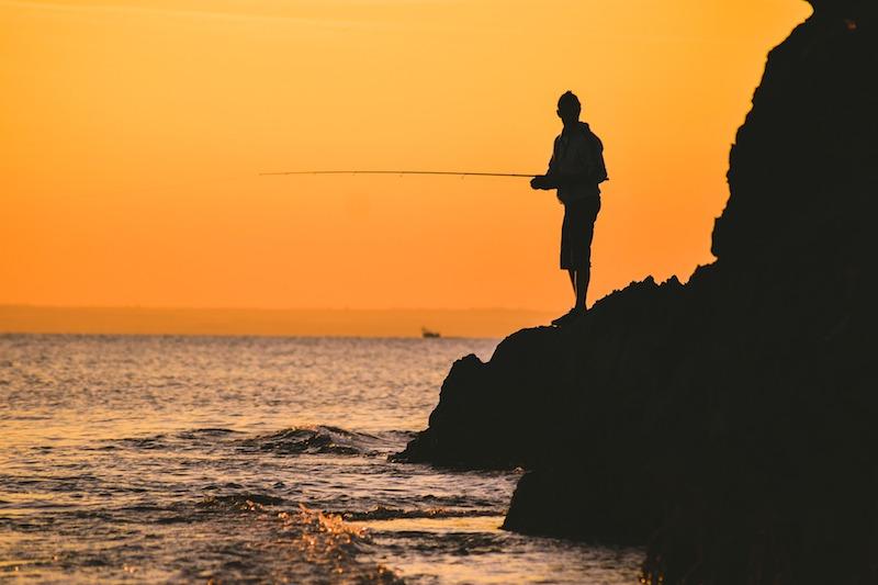 Рыбак с удилищем ловит рыбу с камней на закате