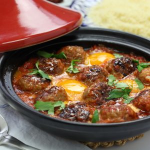 Small Moroccan meatballs and poached eggs in tomato sauce sauce are ready to be served from a tagine. The dish is garnished with fresh parsley and part of the red conical lid can be seen resting against the tagine base.