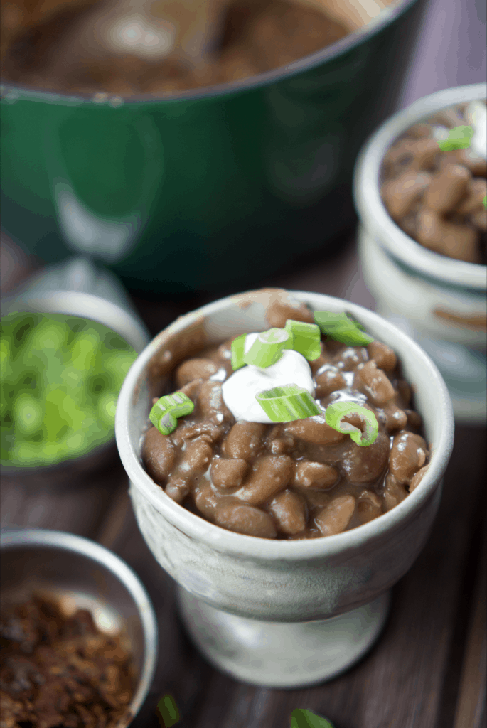 Oven baked pinto beans in ceramic cups surrounded by pot of beans and toppings.