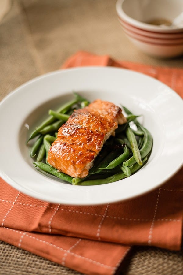 Single fillet of salmon that has been pan fried and is plated over garlic parmesan green beans. The spice rub for the pan fried salmon can be seen in the background.