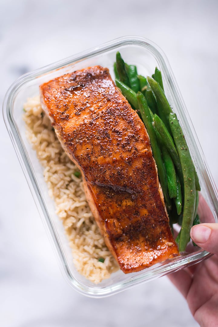 Hand holding a single meal prep container which has pan-roasted salmon, garlic rice, and parmesan garlic green beans for how to meal prep salmon.